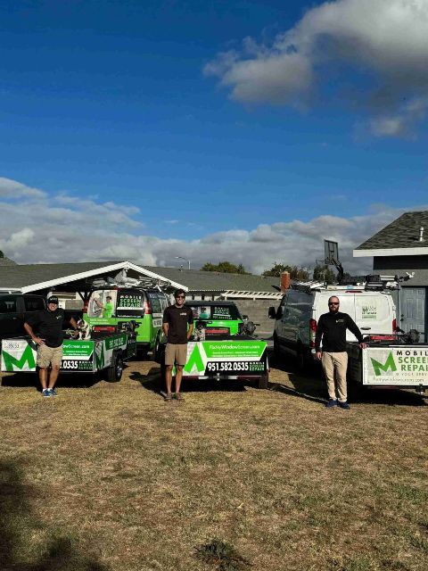 Three men standing near green and white work trucks, in front of a building on a sunny day.