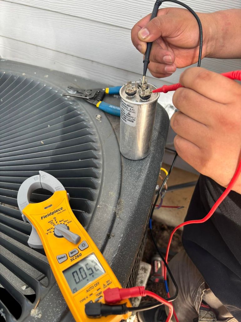 Person using a multimeter to test a cylindrical capacitor on an AC unit; clamp meter nearby displays a reading.