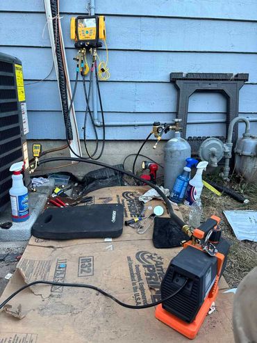 HVAC equipment setup against a blue house: gauges, vacuum pump, tank, tools on cardboard.