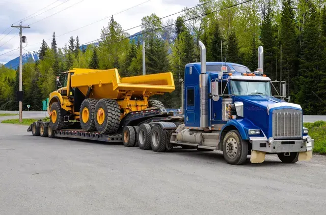 a blue semi truck is carrying a yellow dump truck on a trailer