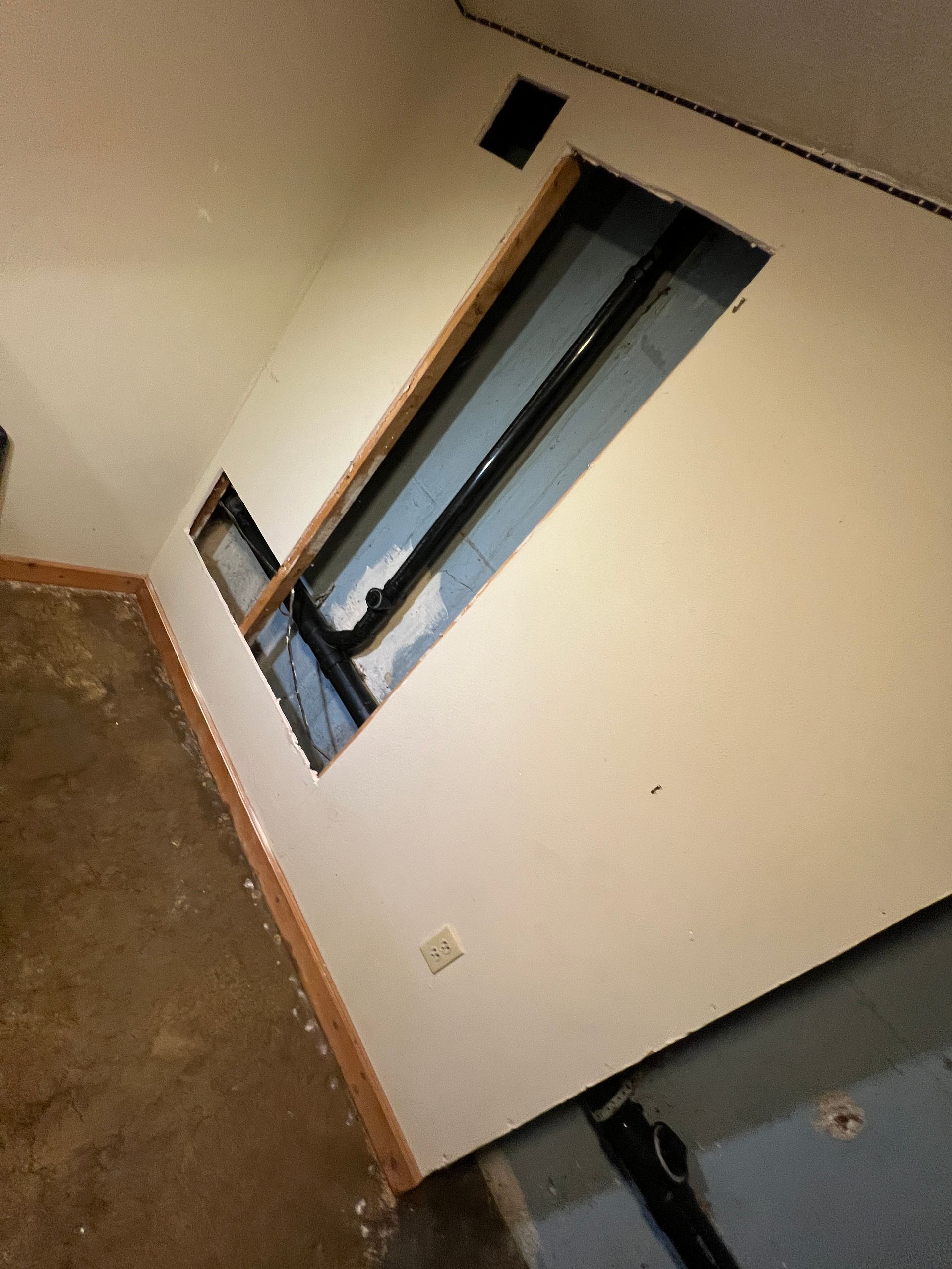 Wall with exposed plumbing in an unfinished room. Brown floor, white wall, black pipes.