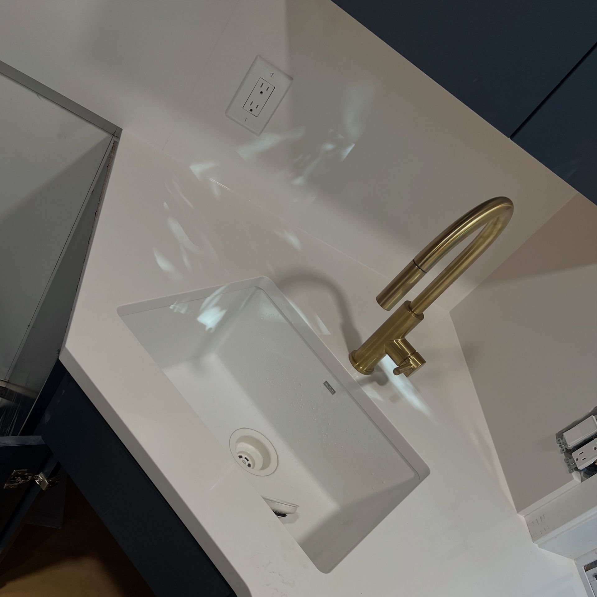 Gold faucet over a white kitchen sink. White countertop with outlet in the background.
