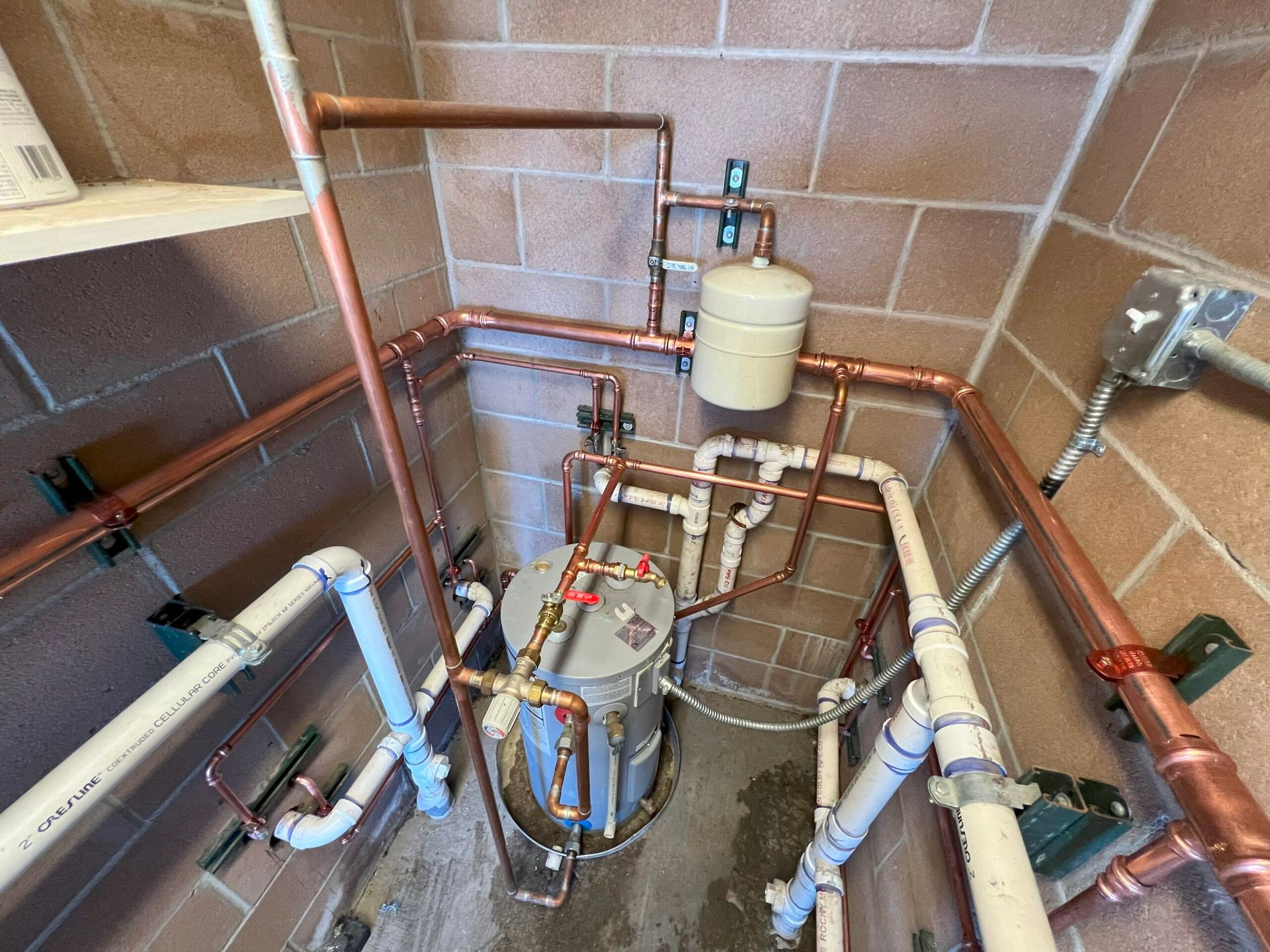 Plumbing system with copper and white pipes, water heater, and expansion tank against brick wall.