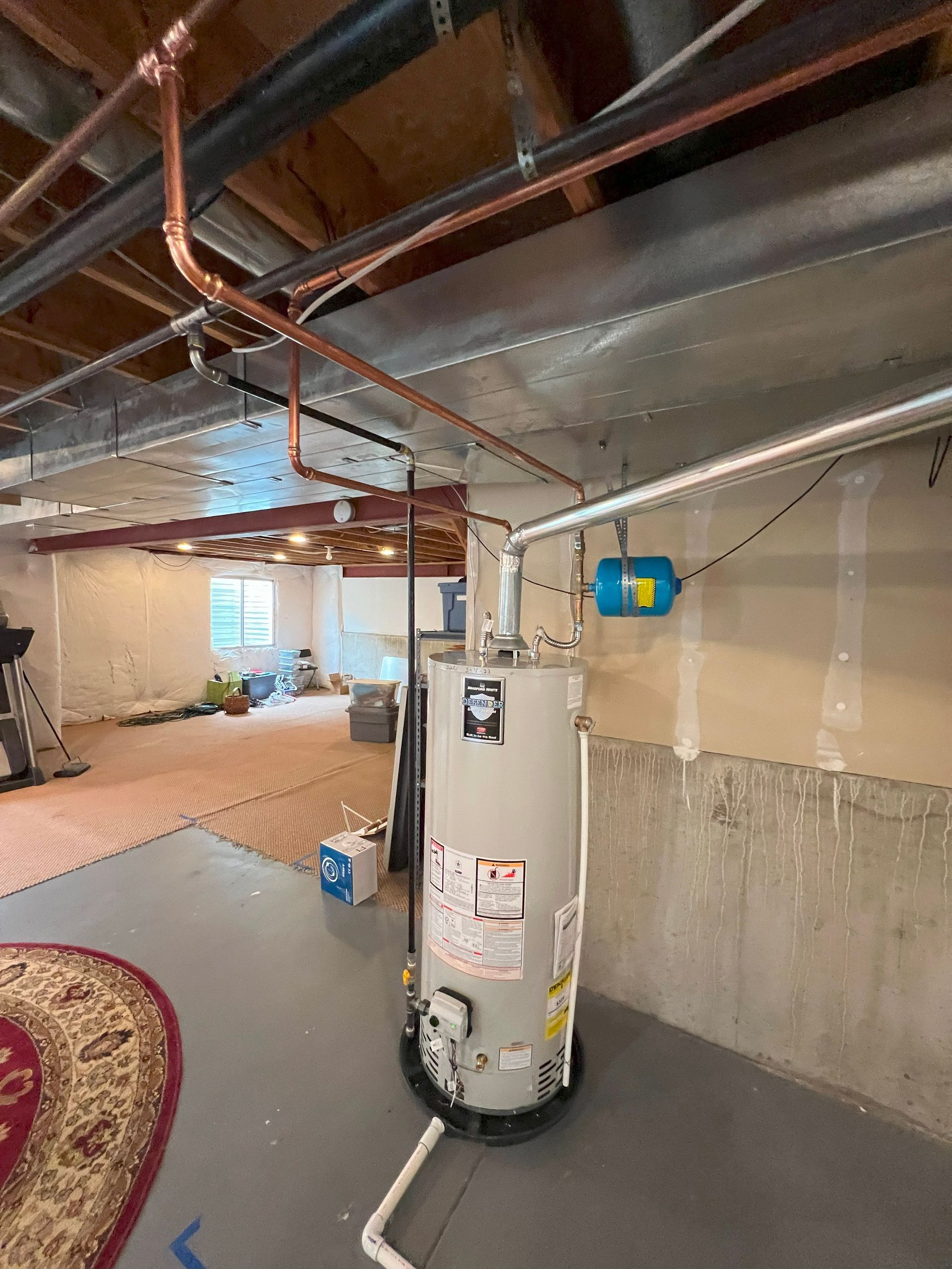 Water heater in a basement, copper pipes overhead, blue expansion tank on wall.