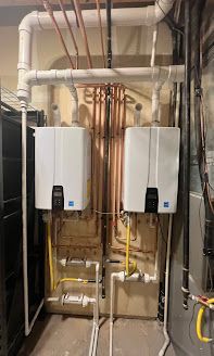 Two white tankless water heaters mounted on a wall with copper pipes and gas lines.