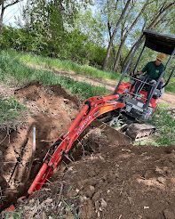 Person operating an orange excavator, digging a trench in the dirt near a grassy bank and trees.
