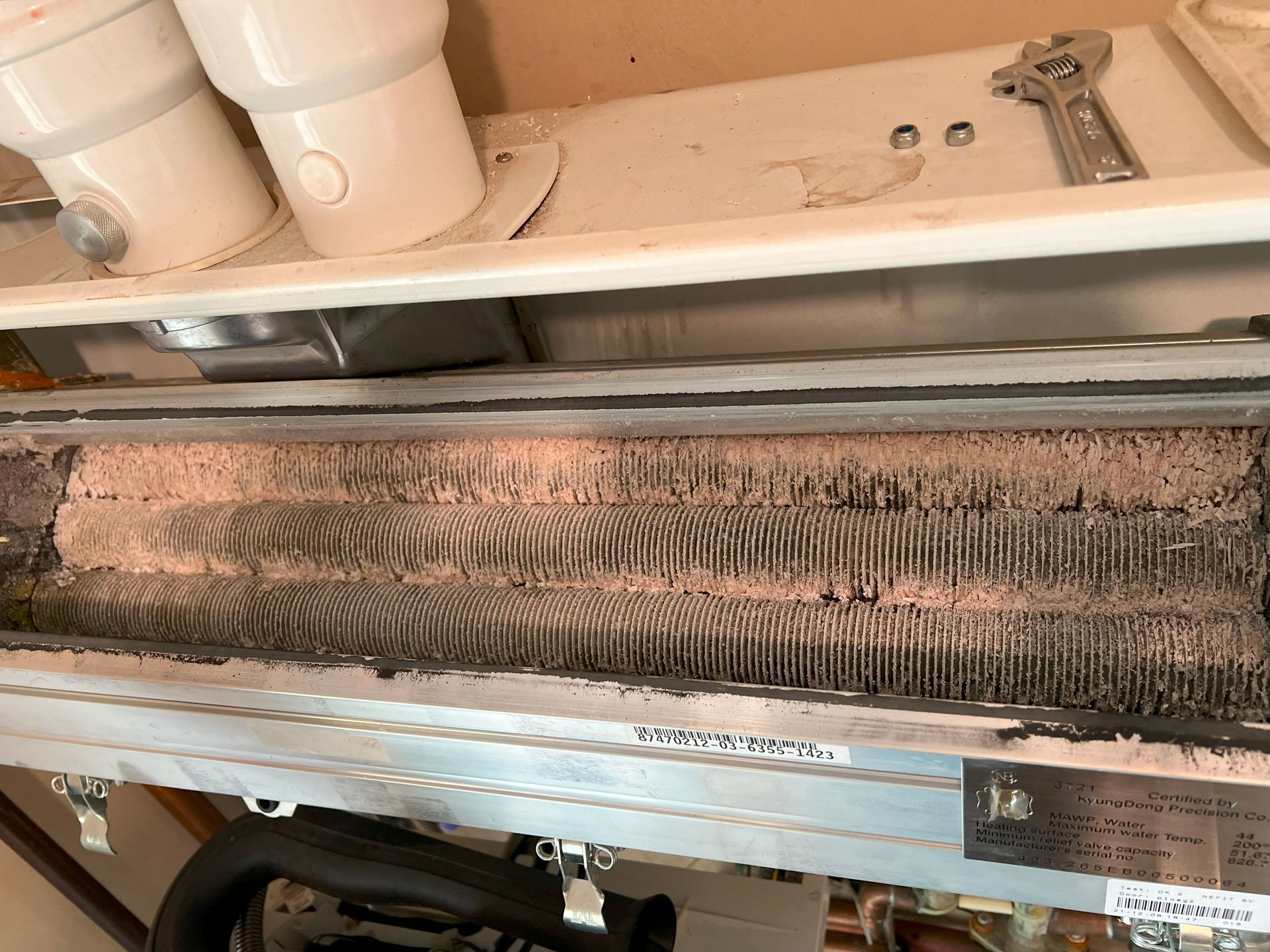 Close-up of a dusty heat exchanger coil with fins, near a white panel and pipes; a wrench is visible.