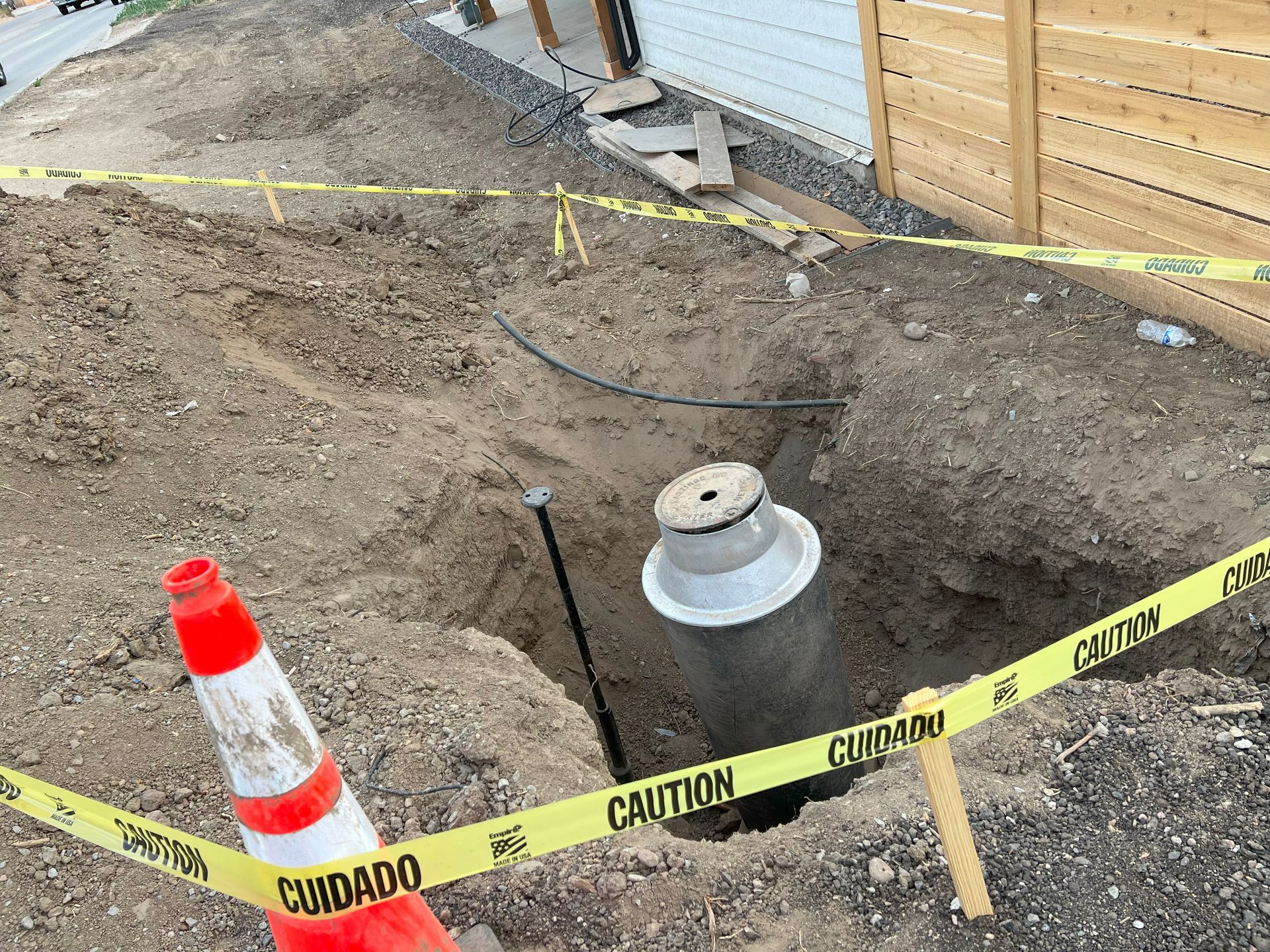Construction site: open hole with a large cylindrical object, caution tape, orange cone, and dirt.