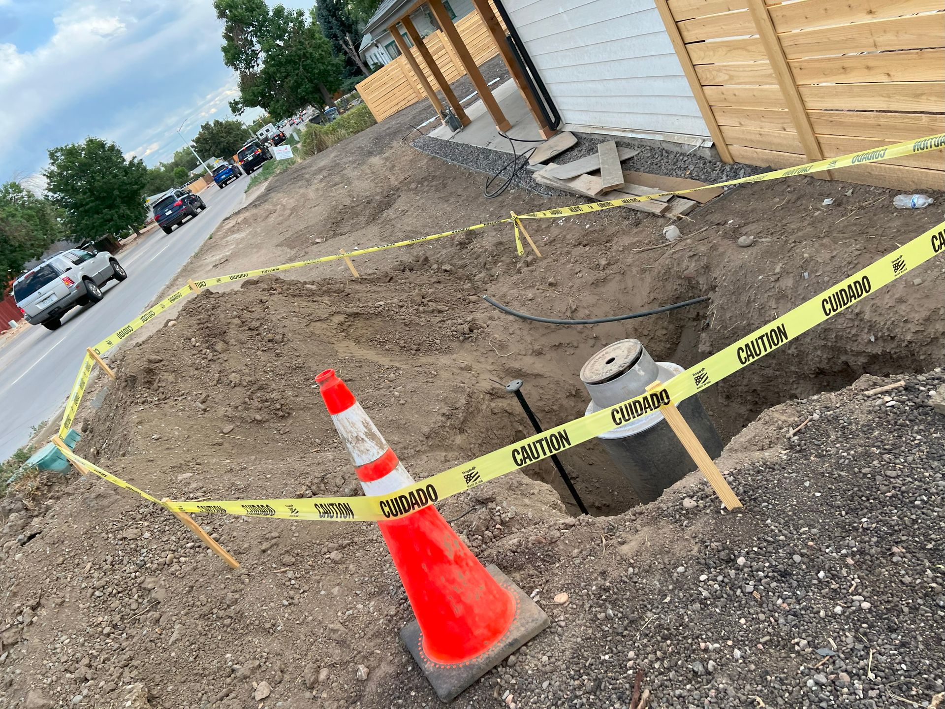 A construction site by a road with a hole, caution tape, and an orange traffic cone.