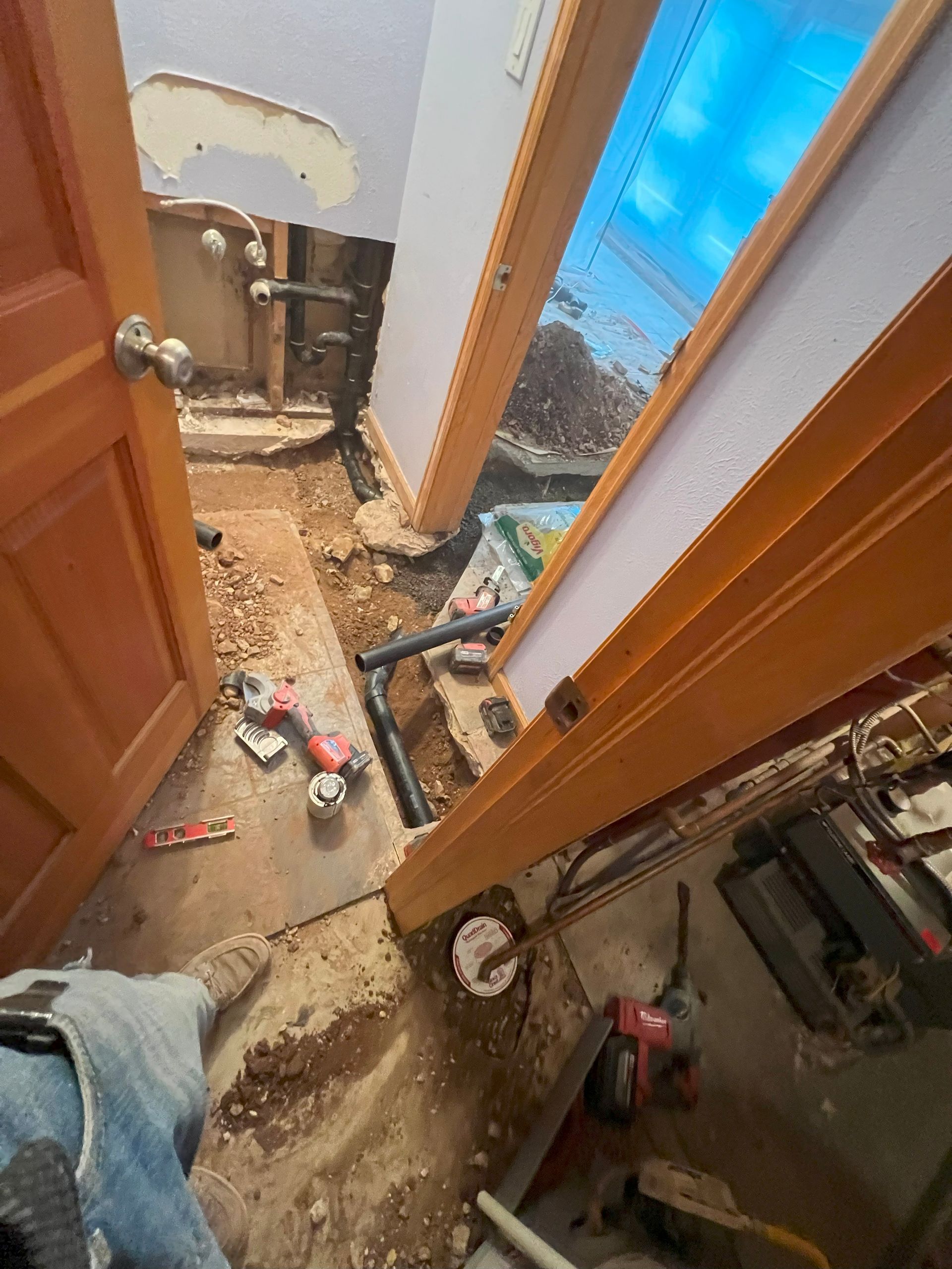 Bathroom under construction: pipes exposed, debris on floor, doorway. Red power tools, muddy area.