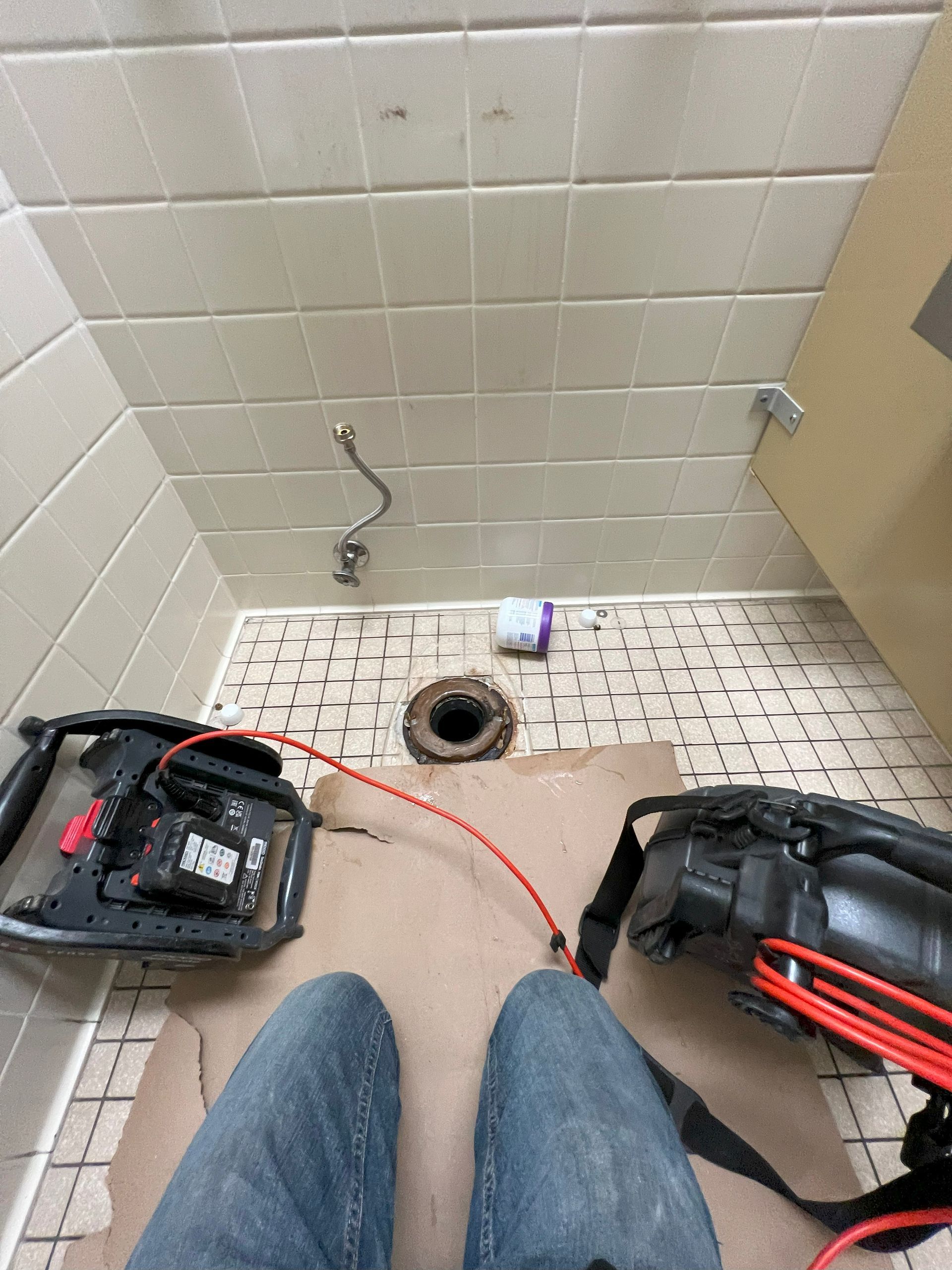 Person in jeans sitting in a shower stall, tools and exposed plumbing visible.