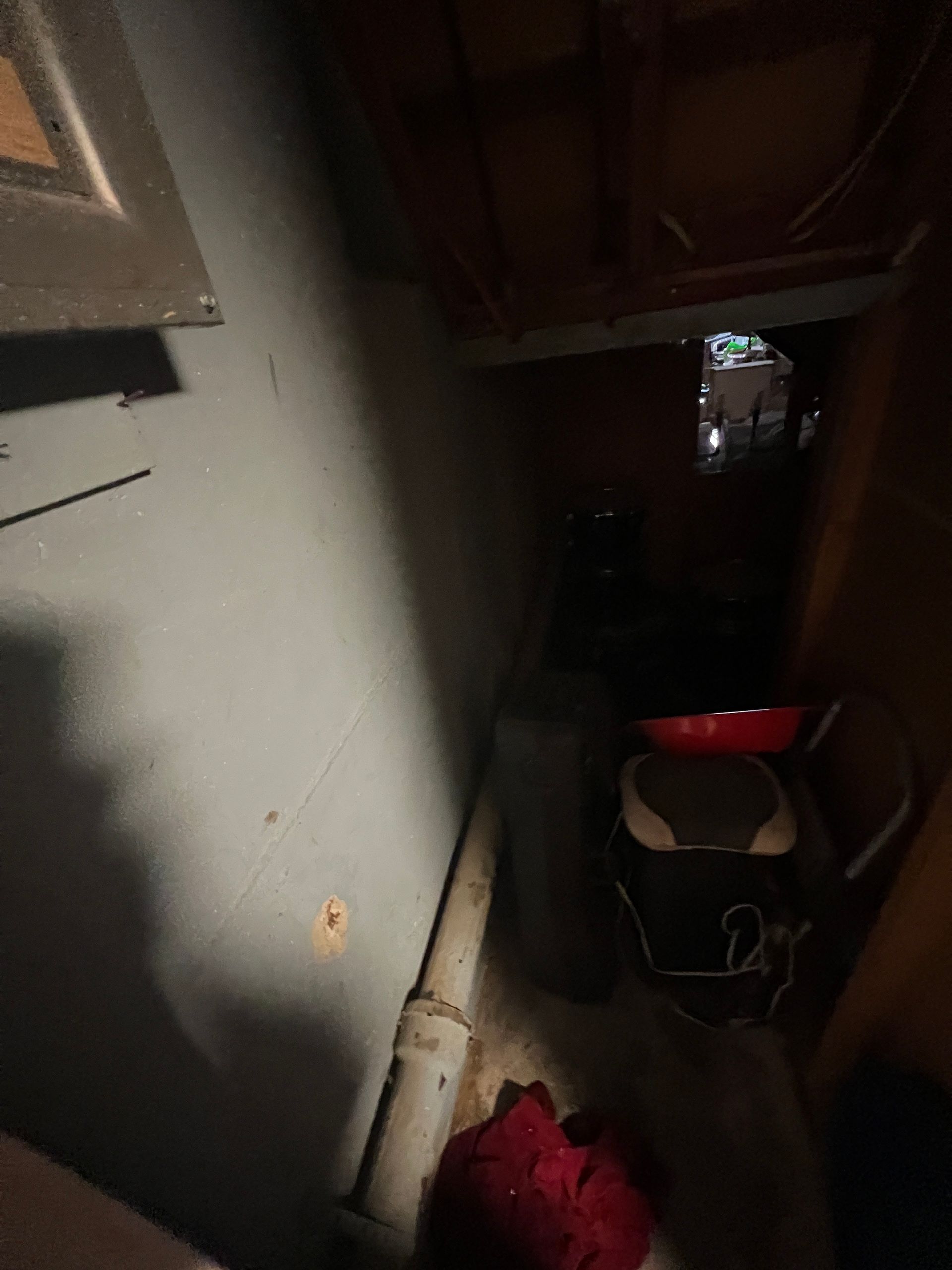 Dark, narrow space with gray wall, pipe, and various objects including a red bowl and small black appliance.
