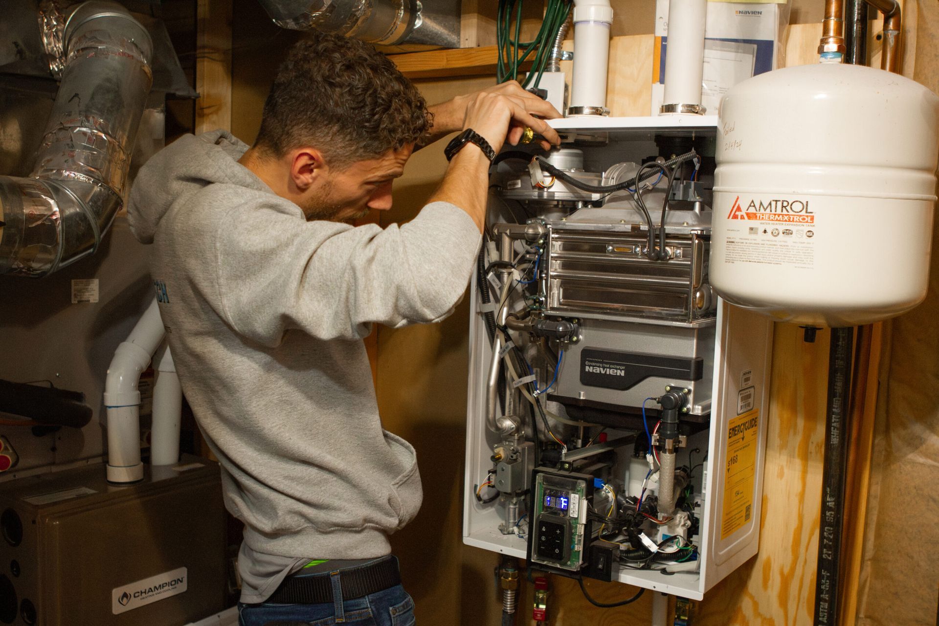 Man in gray hoodie inspecting the interior of a wall-mounted water heater in a utility room.