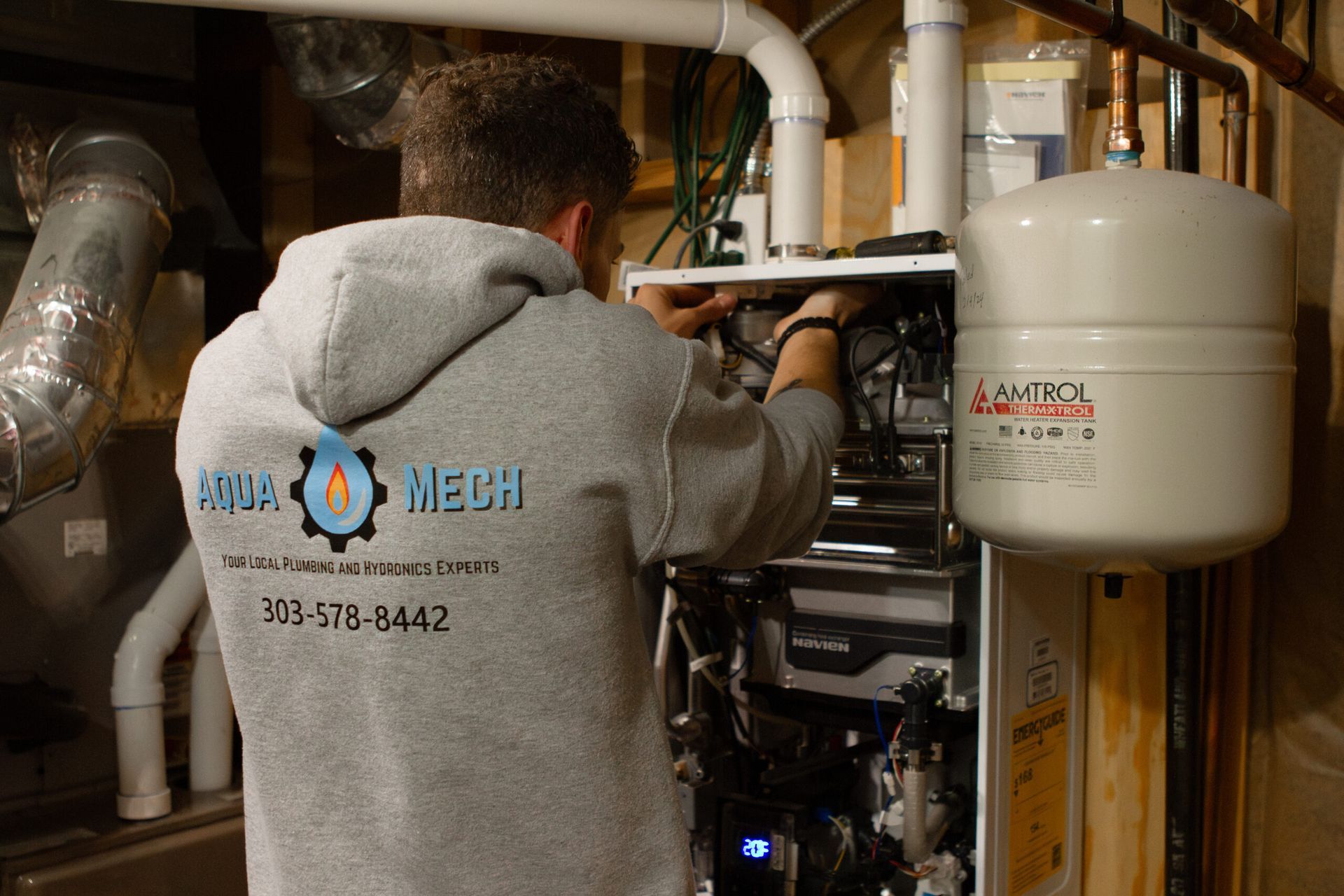 A person in a gray hoodie works on heating equipment in a basement. The hoodie has the text 