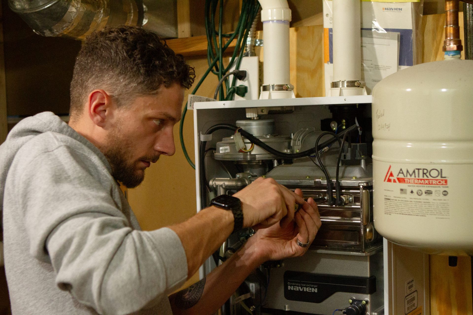 Man working on a heating system. He's using a screwdriver, indoors, with a white tank visible.