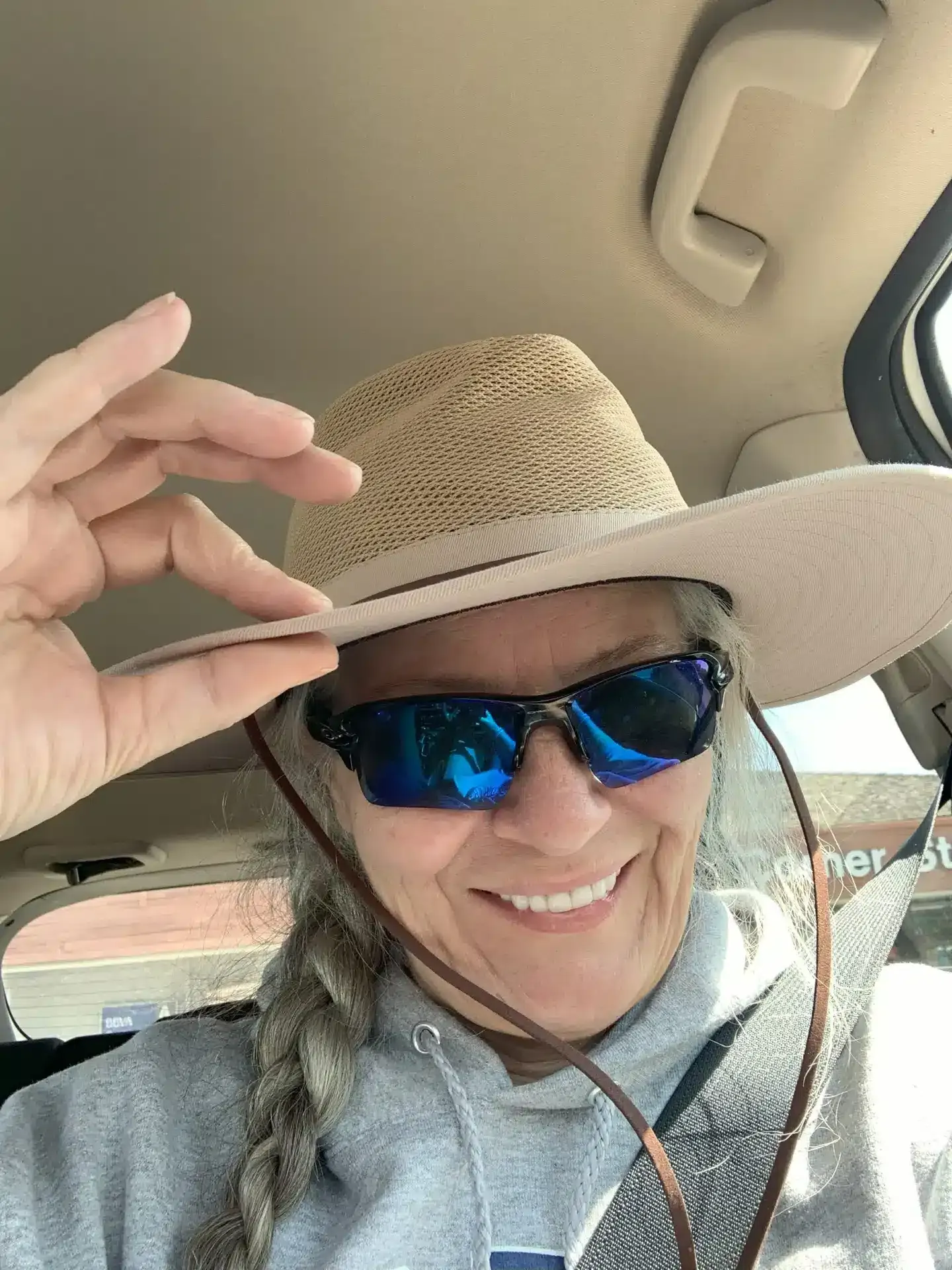 Woman smiling, wearing sunglasses and straw hat, braid, hand touching hat brim, car interior.