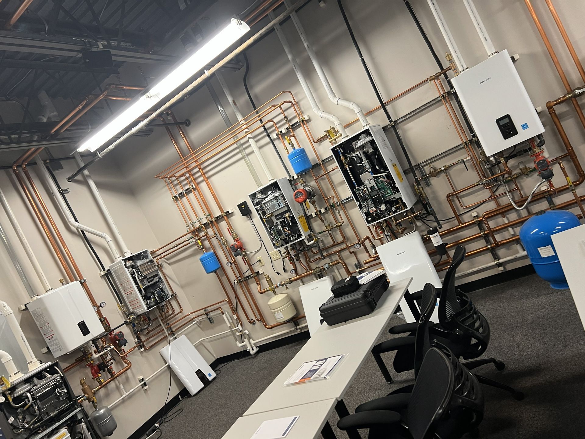 A classroom with several wall-mounted water heaters and copper piping. A table with chairs is in the foreground.