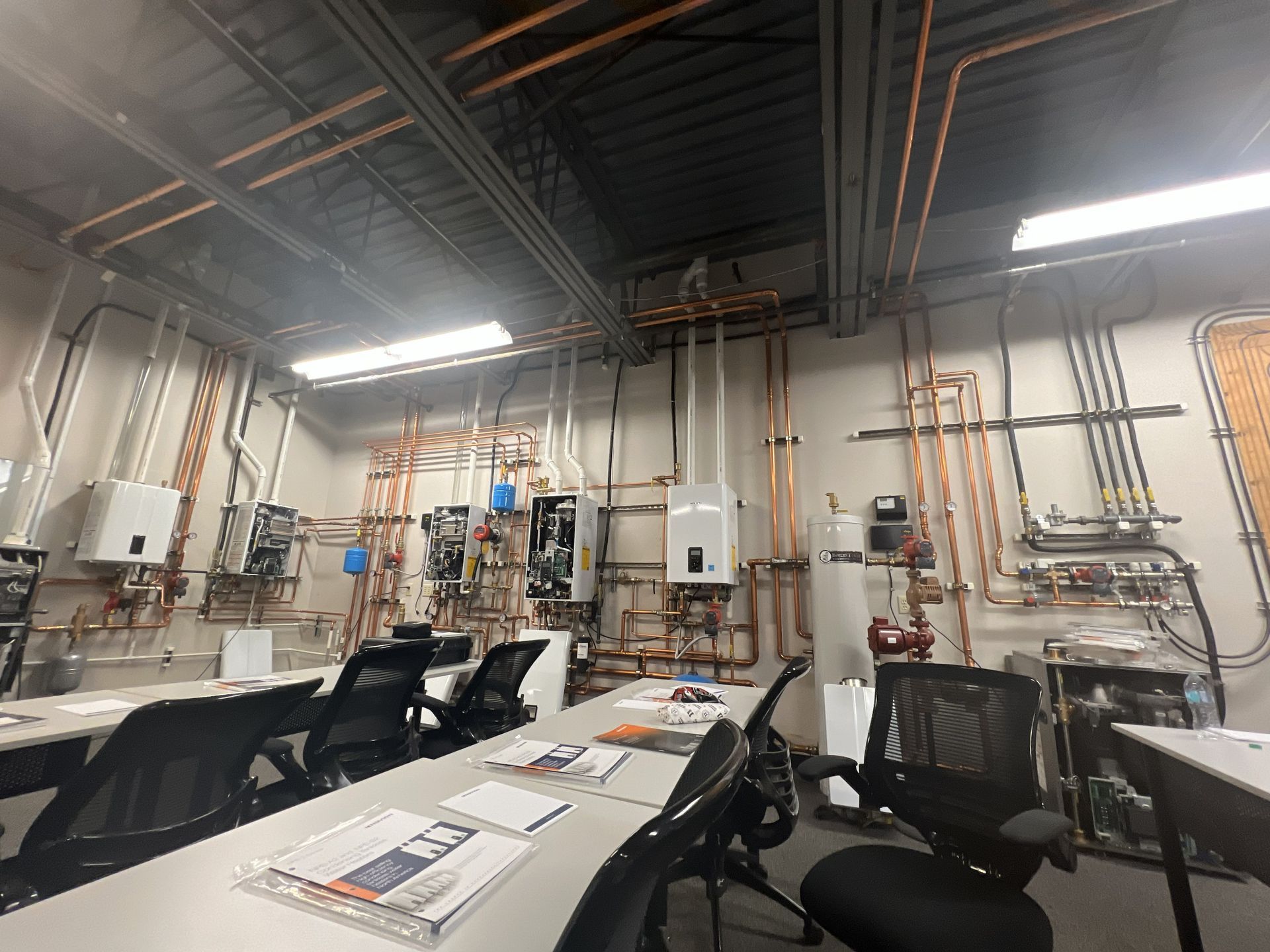 Classroom with tables, chairs, and mounted plumbing equipment. Copper pipes and boilers against white walls.