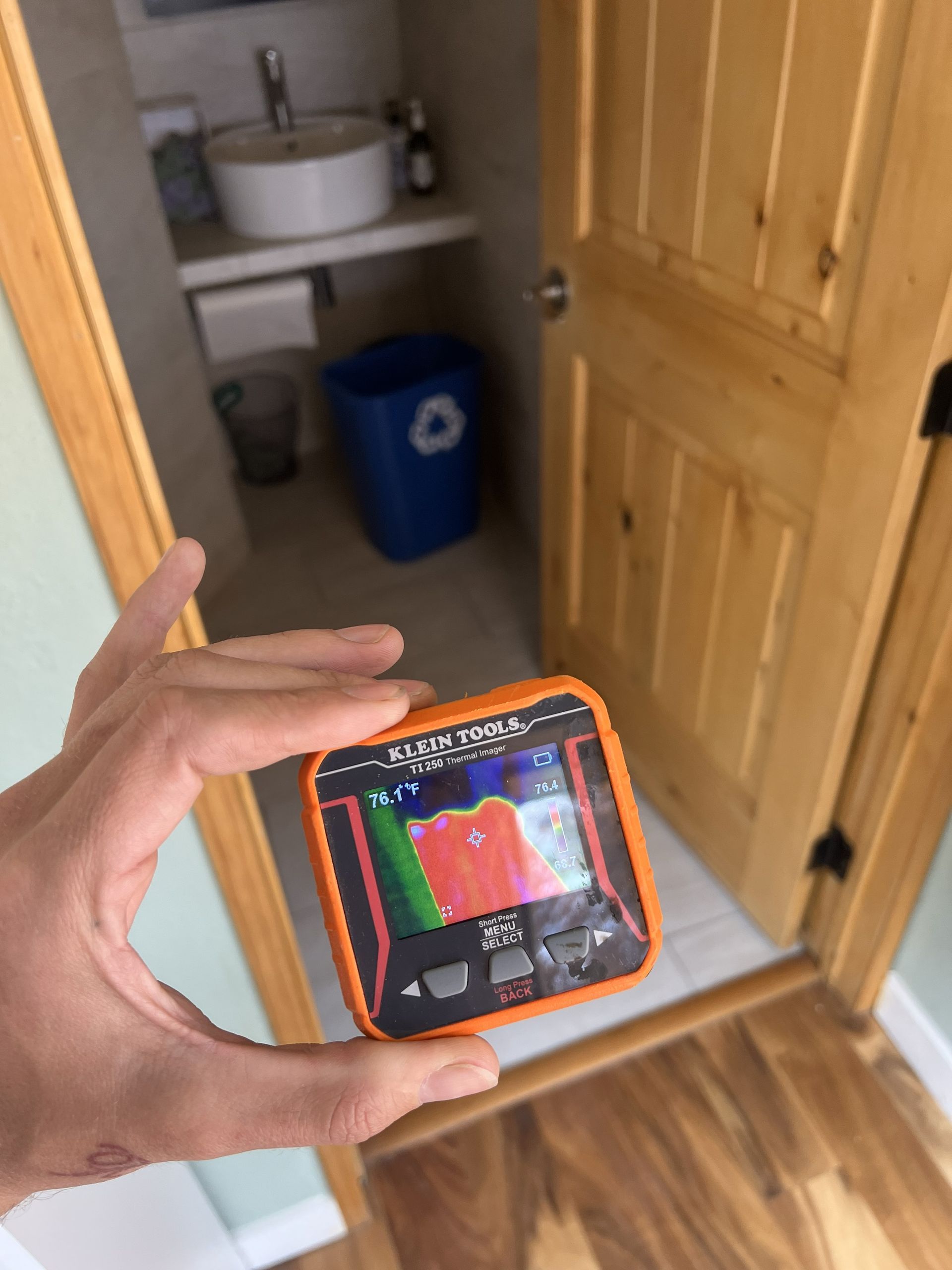 Hand holding a thermal imaging camera pointed at a closed wooden door; a bathroom is visible beyond the door.