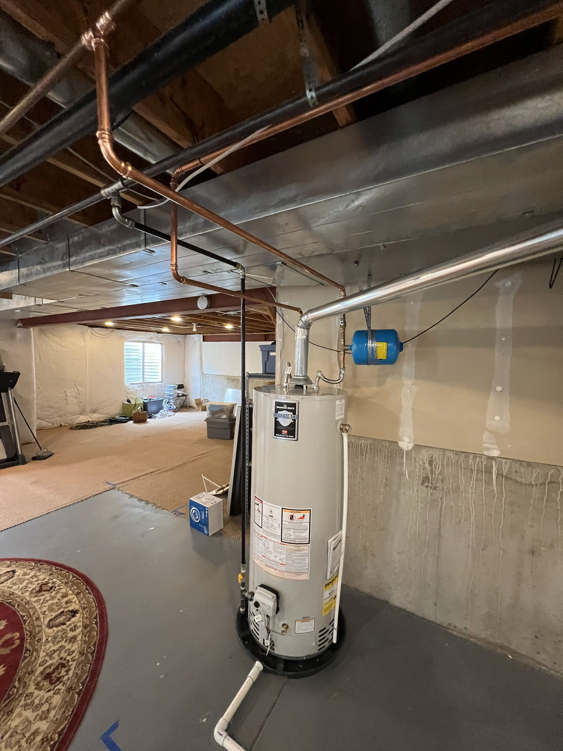 Water heater in a basement with copper pipes overhead. Blue expansion tank on the wall.