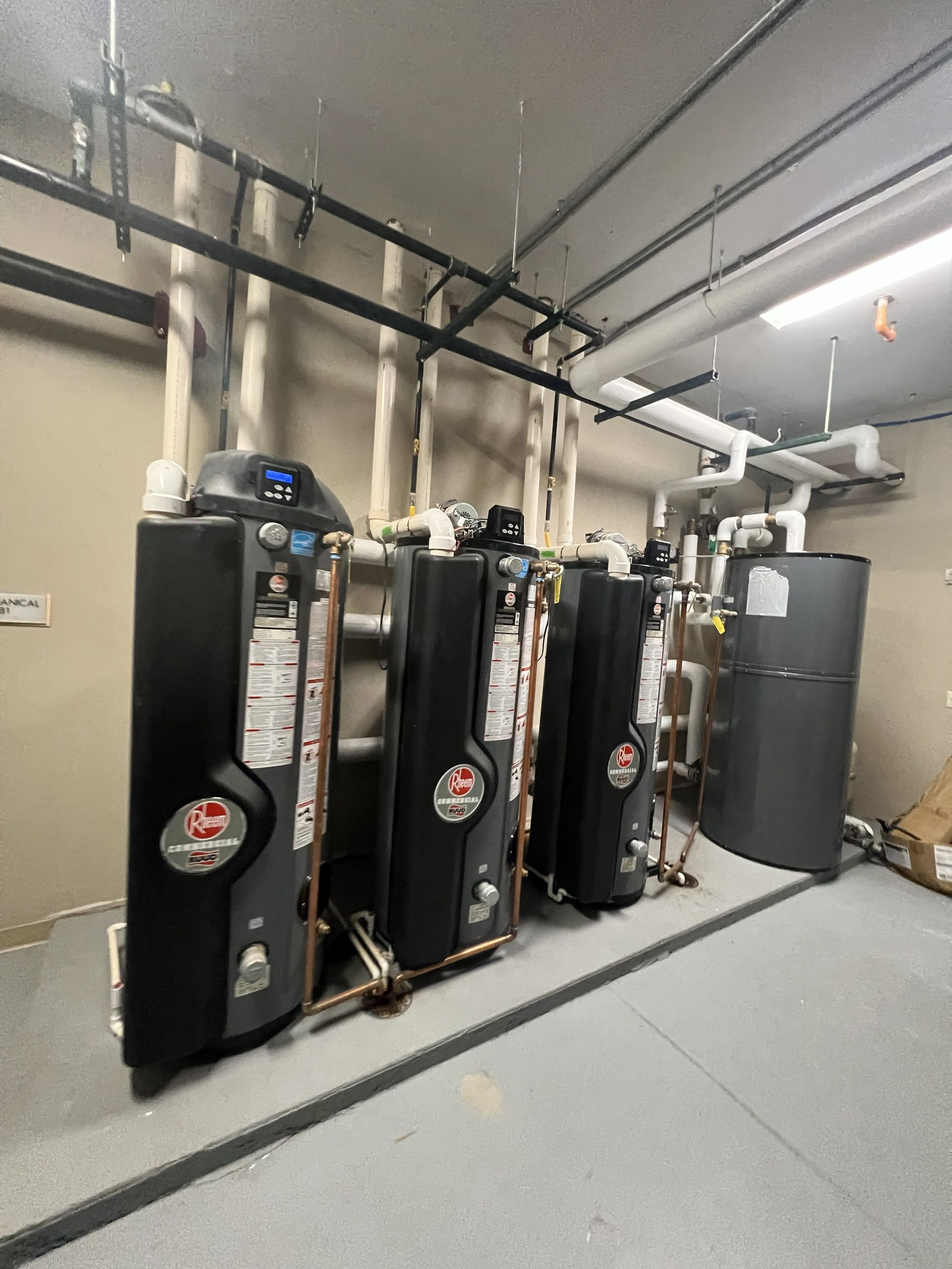 A row of black water heaters with pipes and a storage tank in an equipment room.