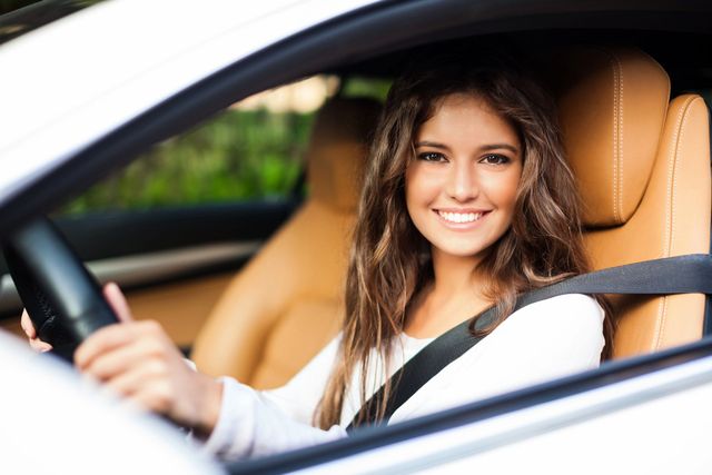Autista sorridente con una maglietta bianca, seduto in un'auto con interni beige e cintura di sicurezza allacciata