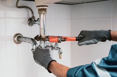 Plumber in gray gloves tightening a sink pipe with an orange wrench under a bathroom sink