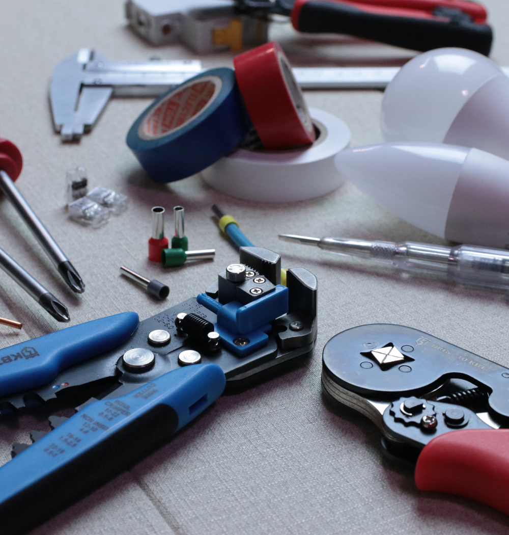 Electrical tools and supplies on a light surface: wire strippers, tape, screwdriver, lightbulb, and crimping tools.