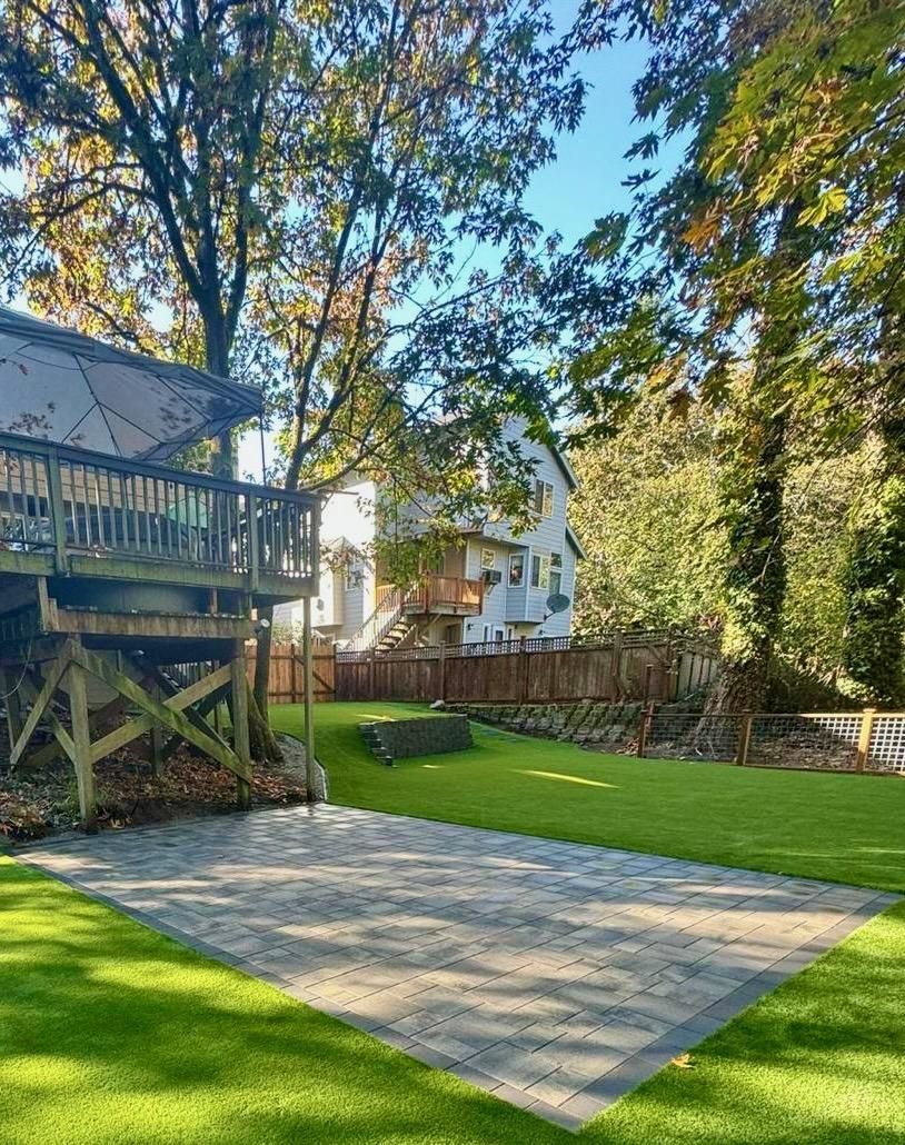Backyard with green lawn, large trees, gravel patio, and a wooden deck attached to a house.