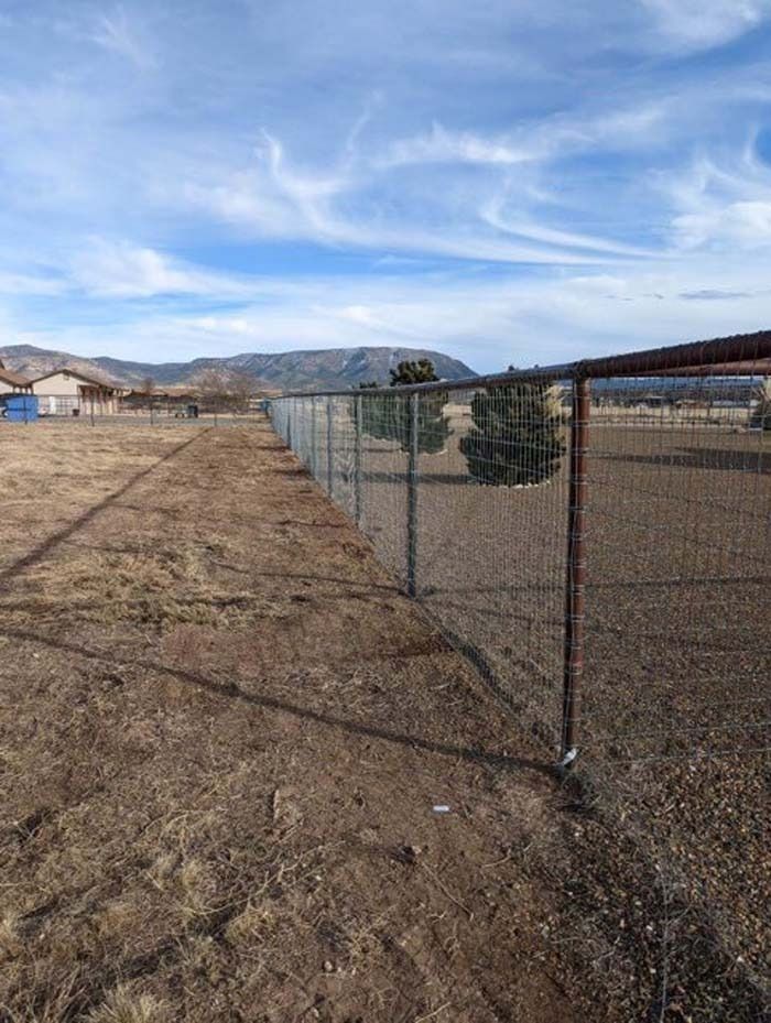 Chain-link fence beside a dry field under a blue sky with wispy clouds and distant mountains