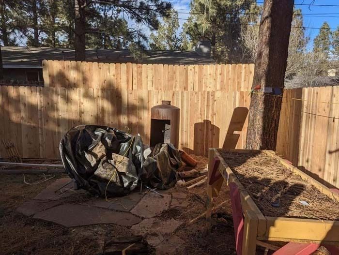 Backyard with a wooden fence, fire pit, and a yellow cart filled with dirt in the foreground.