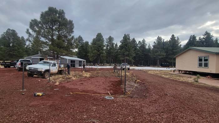 Rural lot with red dirt, parked truck and trailer, small building, and trees under a cloudy sky