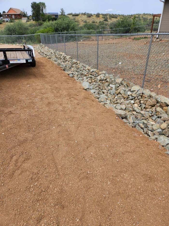 Gravel path beside a chain-link fence and rock border in a rural yard with a trailer at left.