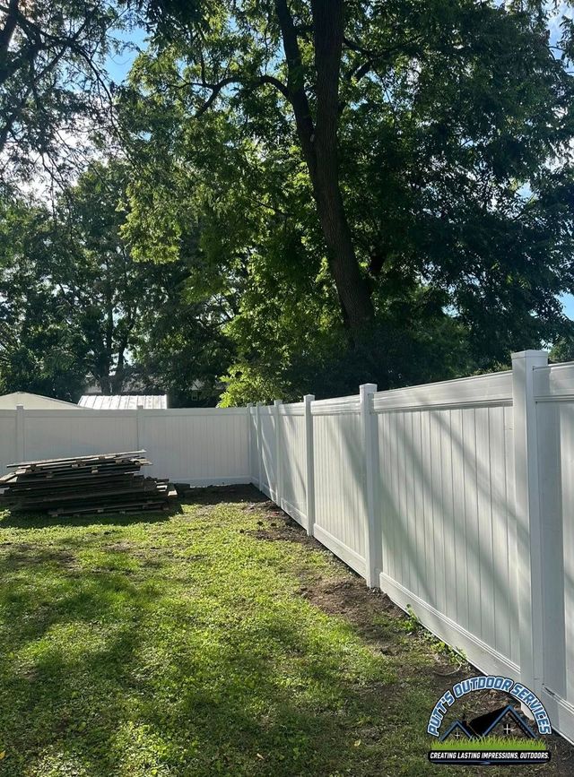 Sunny backyard with white vinyl fence, tall trees, and stacked boards on the grass