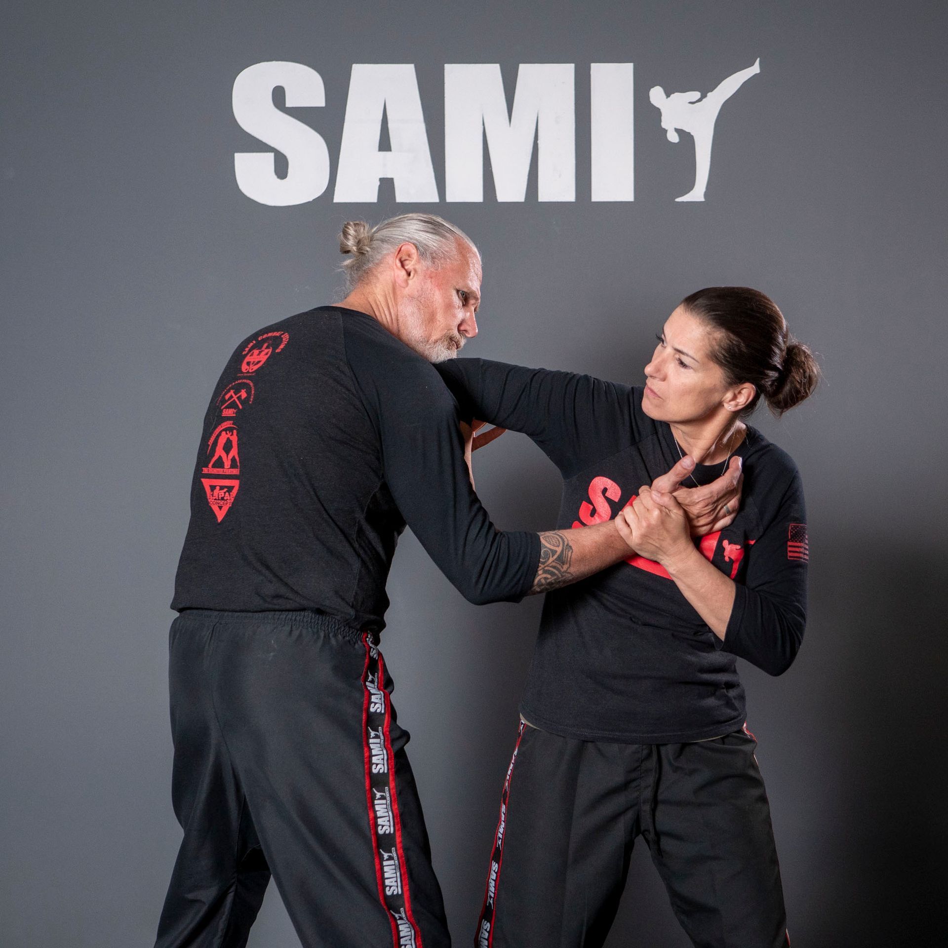 Two men in a grappling hold during a training session. One has a headlock while the other is standing. SAMI logo visible.