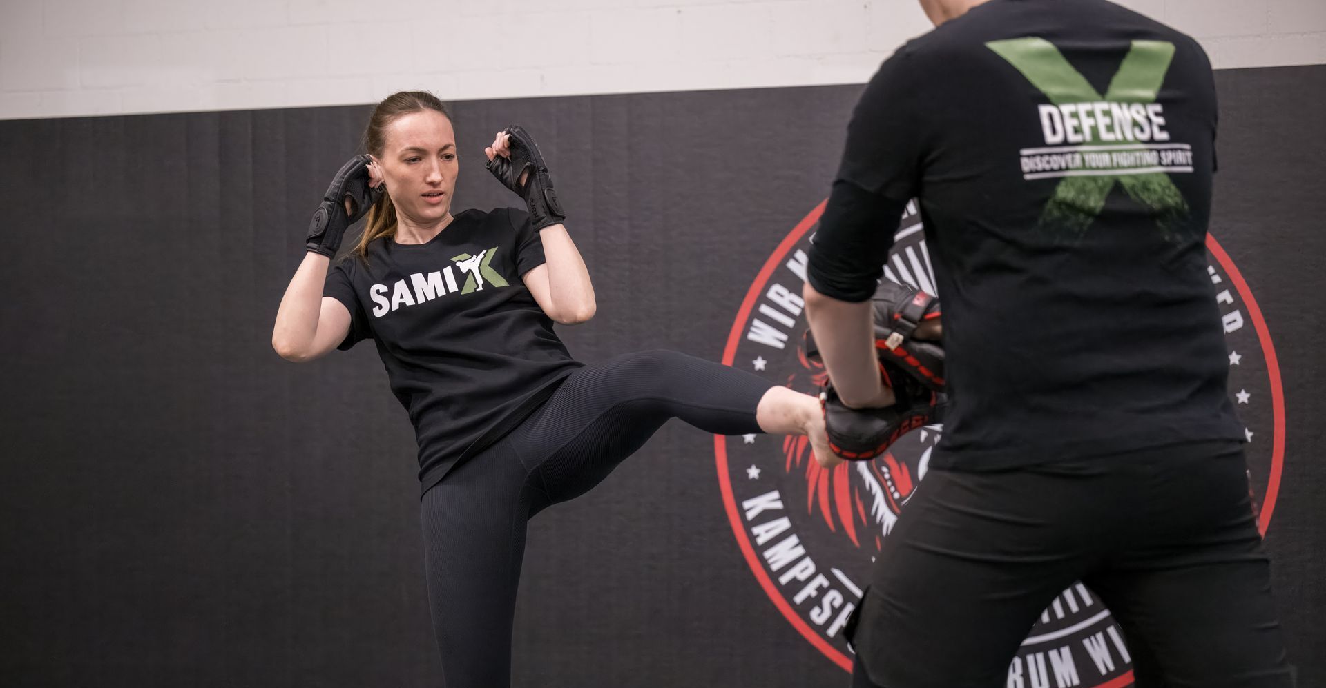 Two men in a grappling hold during a training session. One has a headlock while the other is standing. SAMI logo visible.