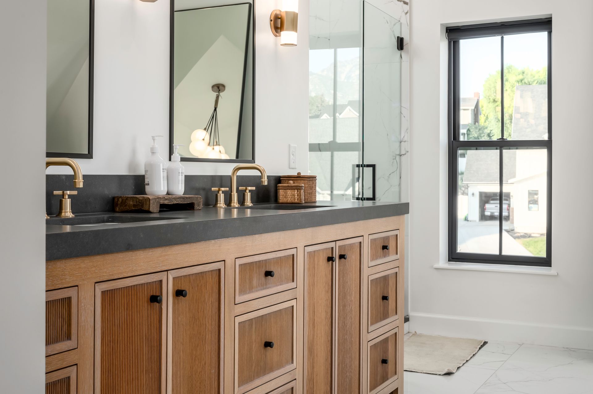 Bathroom with wooden vanity, dark countertop, gold fixtures, large mirror, and window.