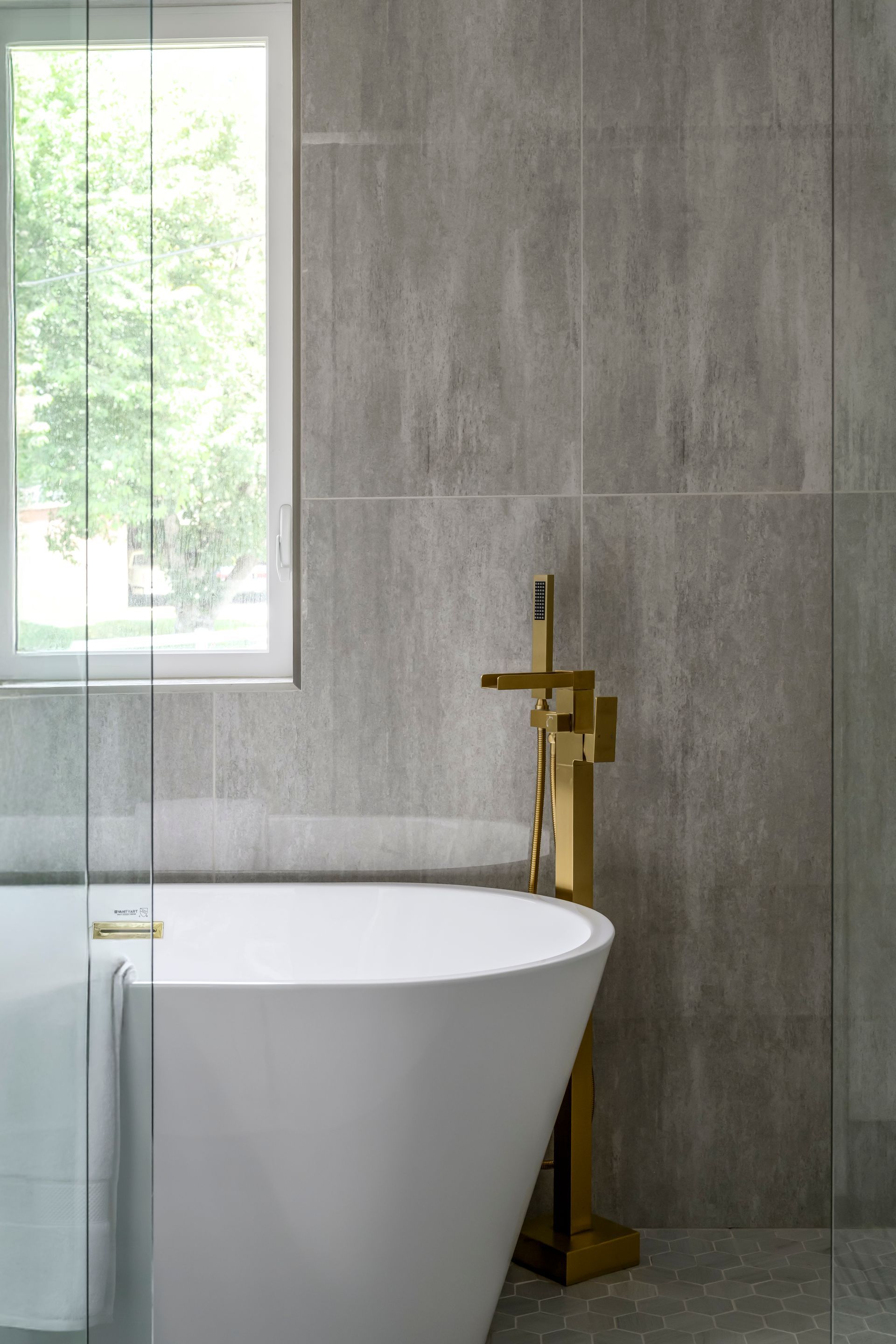 White freestanding bathtub with gold faucet, set against gray tiled wall near a window.