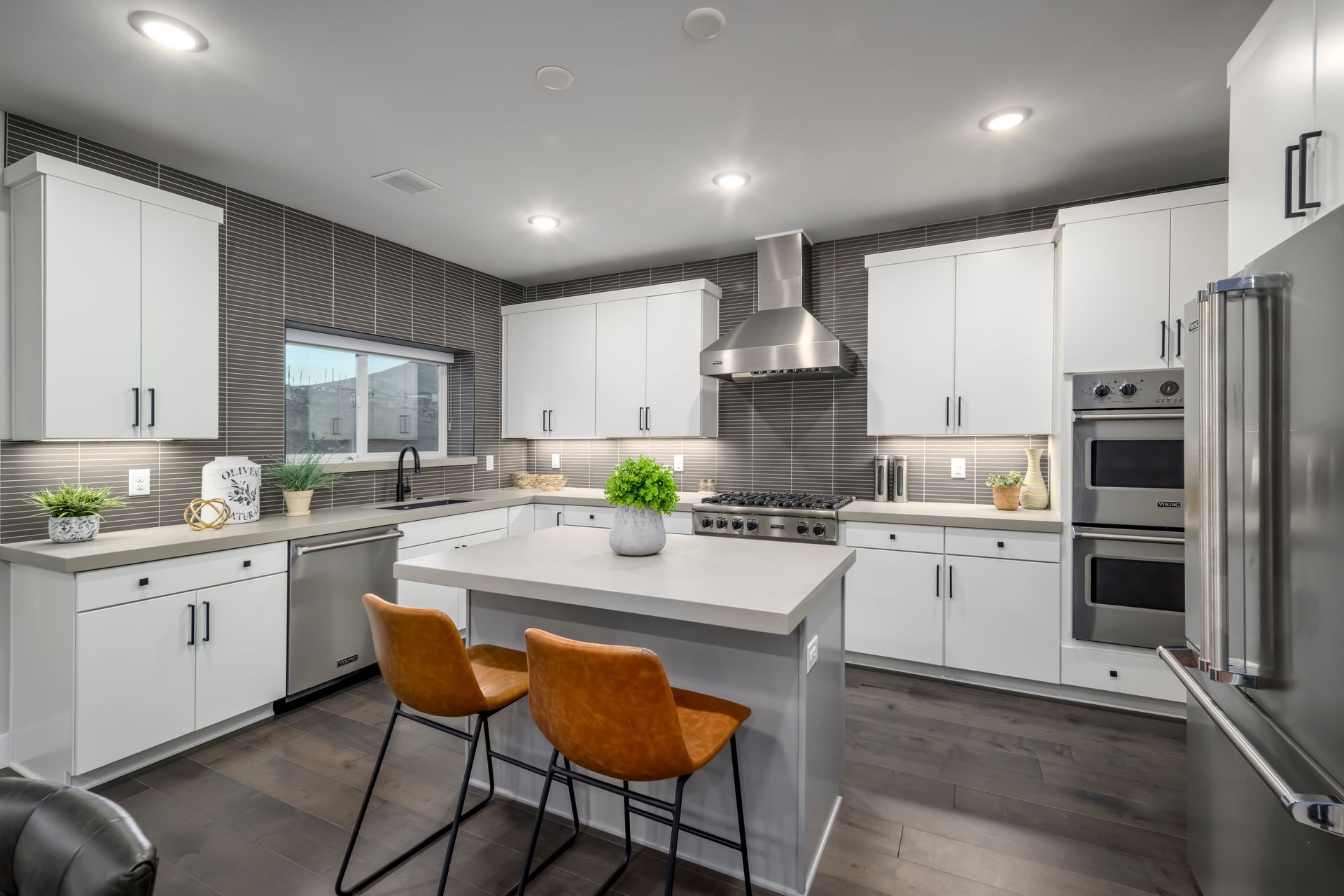 Modern white kitchen with stainless steel appliances, grey backsplash, and a breakfast bar with two brown stools.