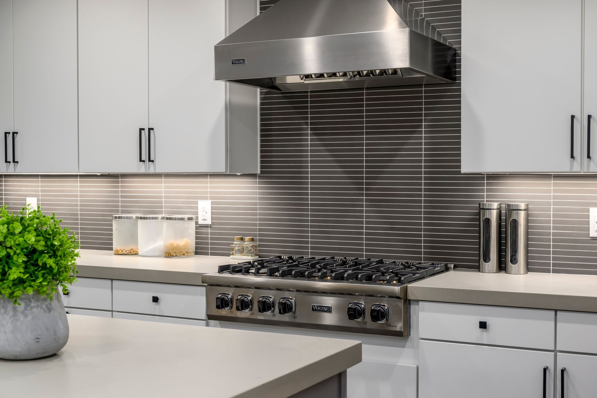 Modern kitchen with stainless steel range, hood, and gray-tiled backsplash. White cabinets, quartz countertop.
