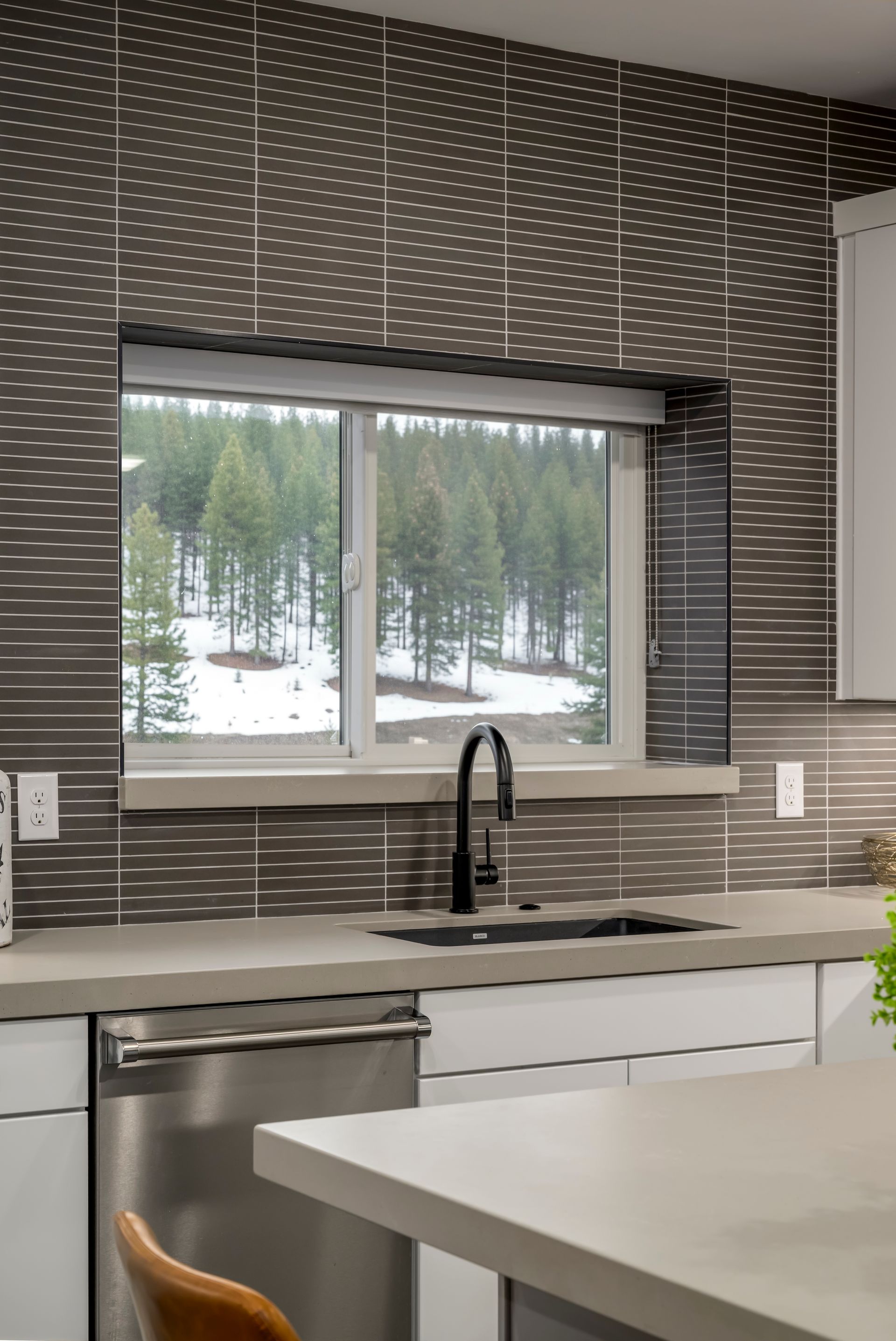 Kitchen with dark patterned tile backsplash, window with snowy view, black faucet, and white cabinets.
