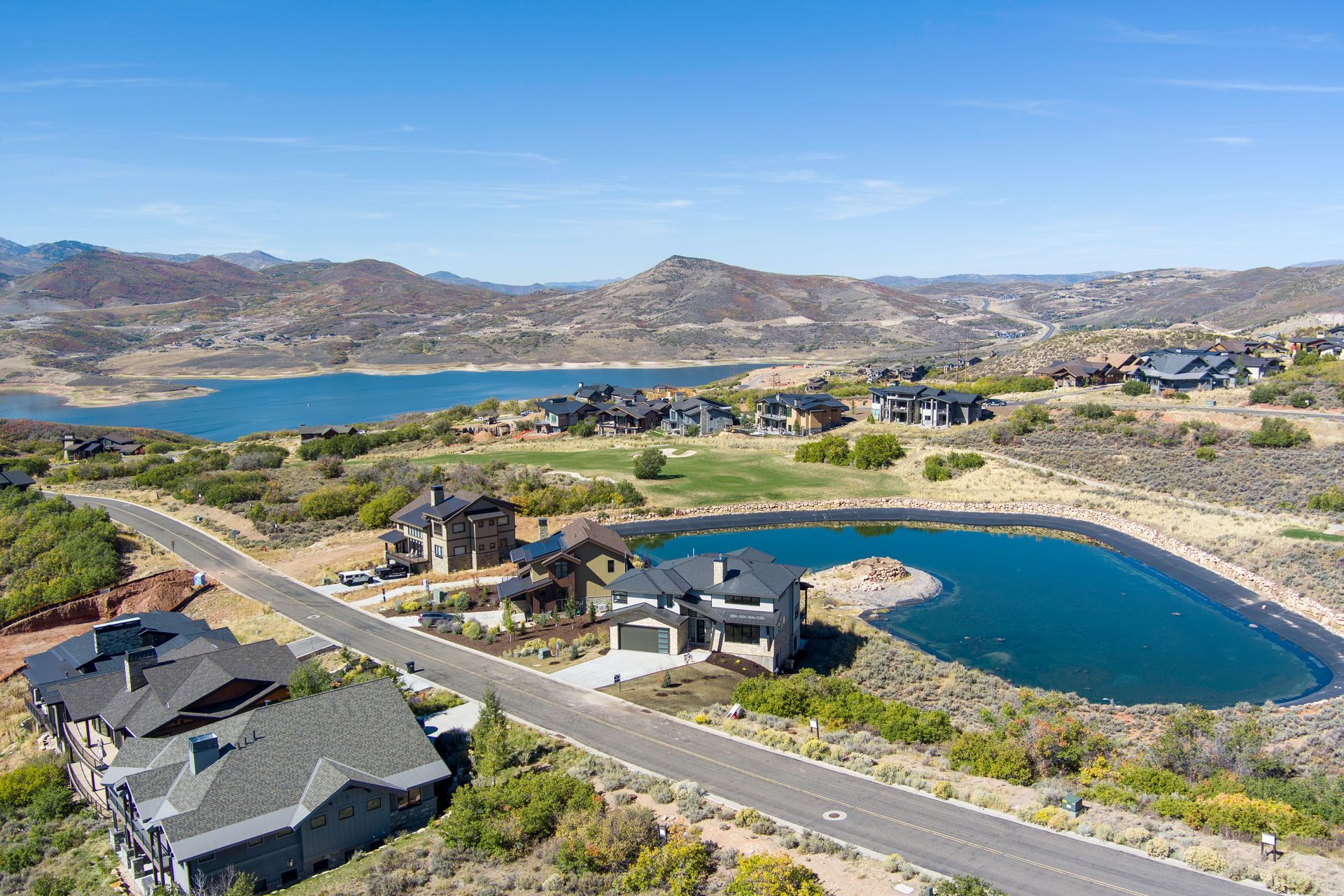 Aerial view of luxury homes near a lake and mountains, blue sky, and winding road.