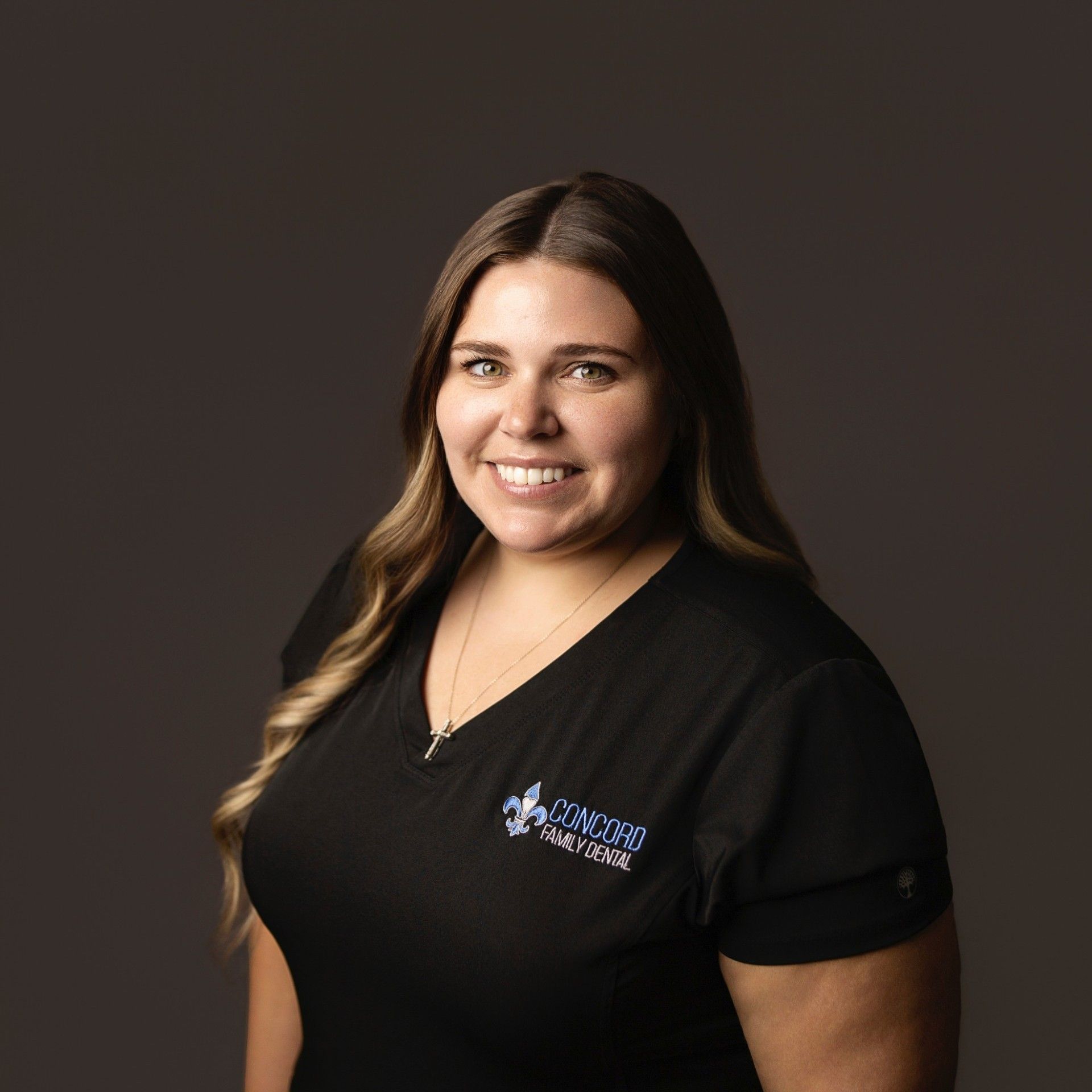 Woman in black scrubs smiling, wearing a necklace. The shirt says