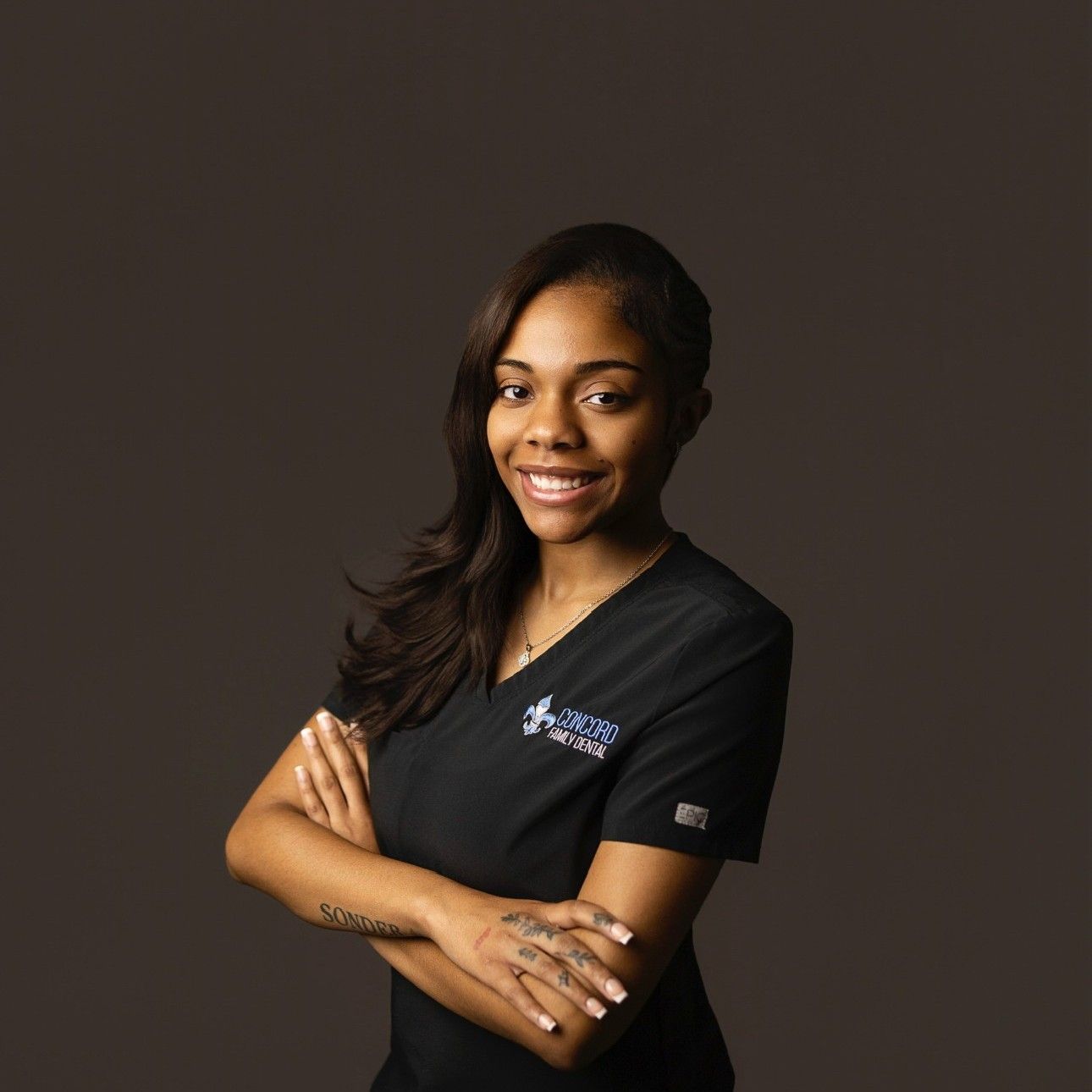Woman in black scrubs, arms crossed, smiling, against a dark gray backdrop.