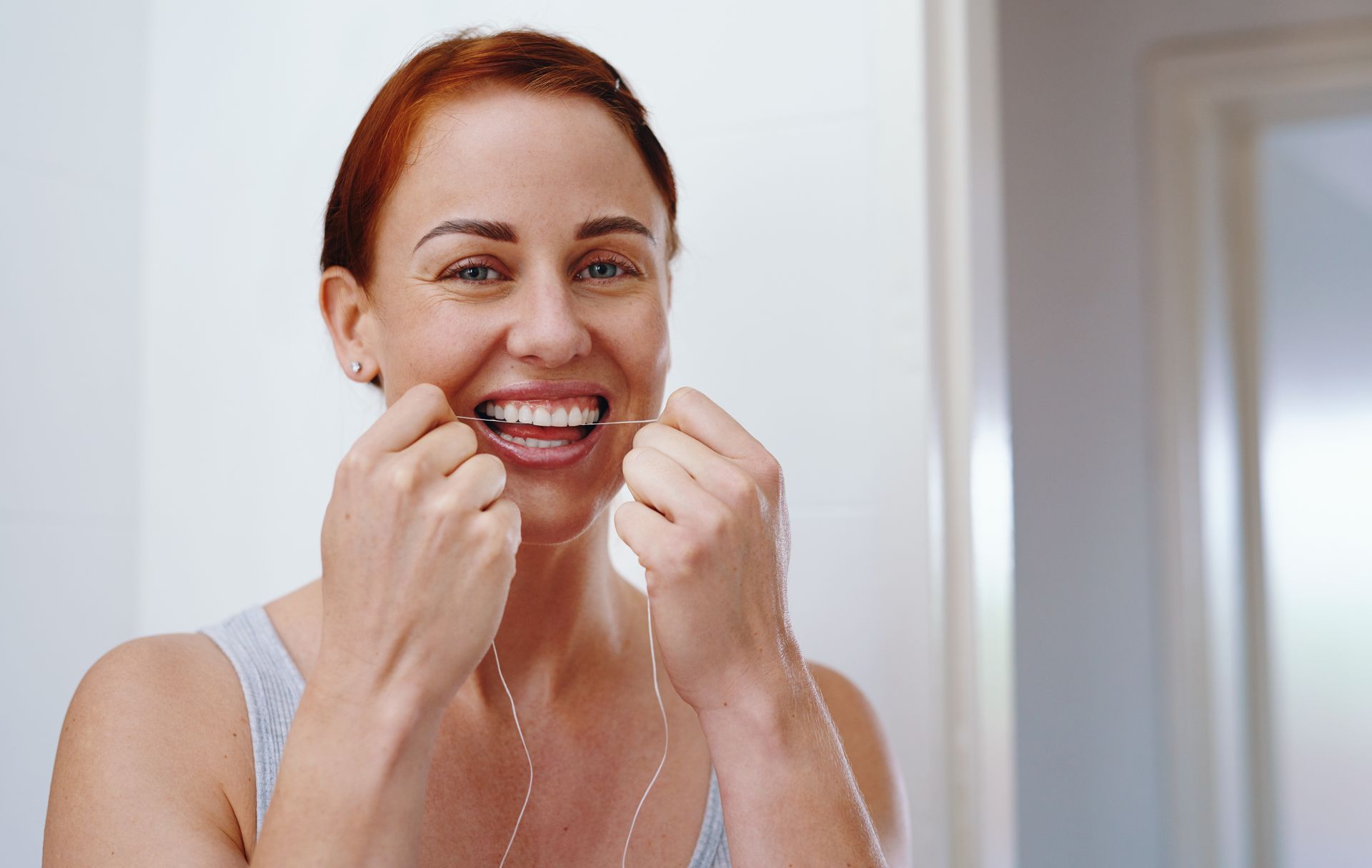 Woman smiling while flossing her teeth in a bathroom