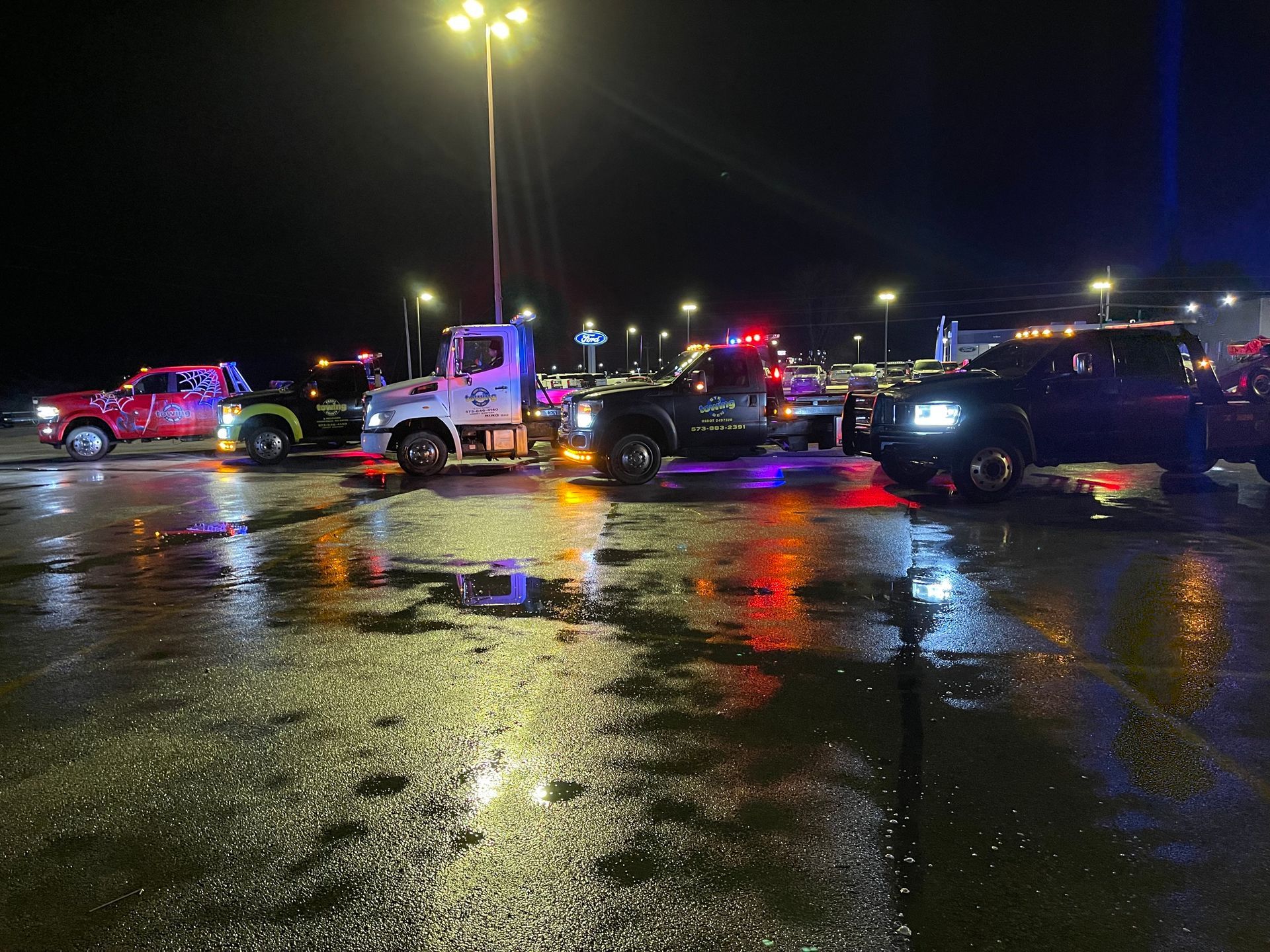 A group of tow trucks are parked in a parking lot at night.