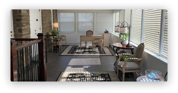 A sunroom with stone walls, striped sunlight, a desk, chairs, rugs, and side tables with lamps.