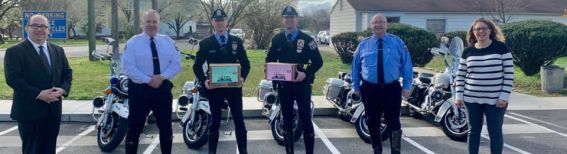 Six people stand in a line outdoors in a parking lot, some holding awards, with several police motorcycles parked behind.