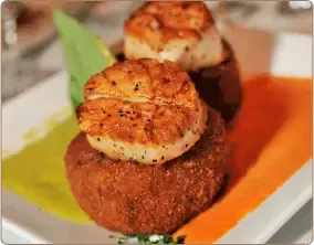 Two seared scallops, each served atop a crispy, round fried cake, drizzled with green and orange sauces on a white plate.