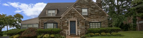 A two-story stone house with a steep roof, central entrance, and bay window, surrounded by green landscaping and trees.